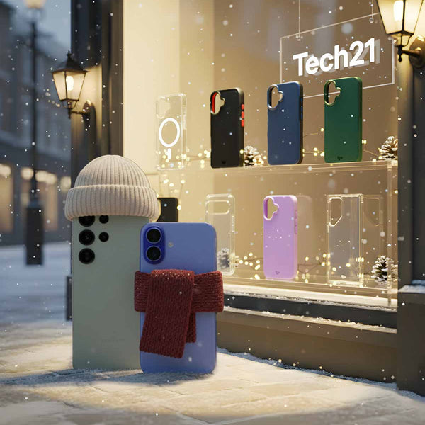 A Samsung S25 and iPhone 17 stand bundled up in knit hat and scarf out in the snow on a street corner looking into a store window with Tech21 cases. Text in the window reads Tech21.